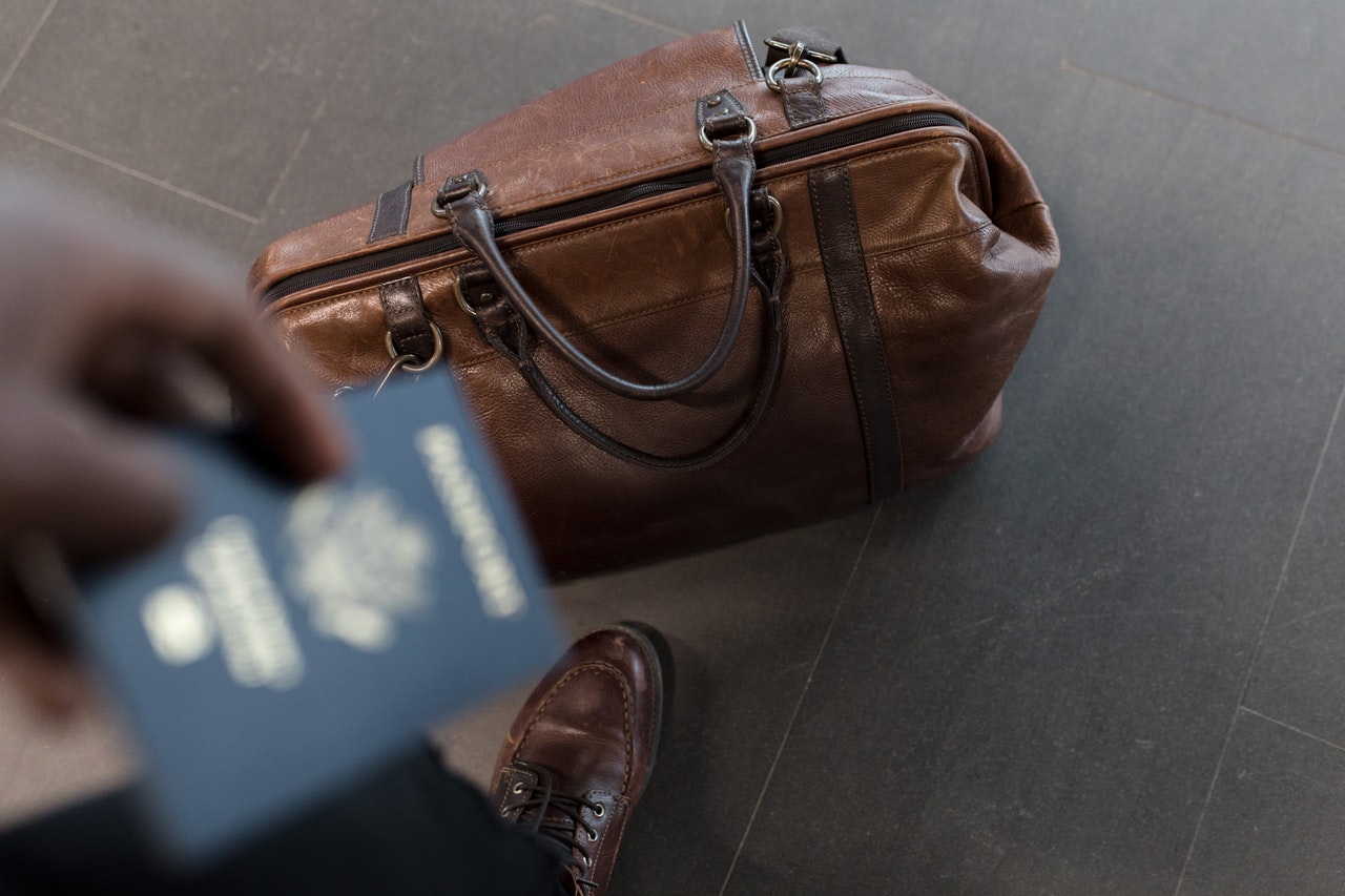 Mężczyzna wyjeżdza w delegacje. Trzyma w dłoni paszport a obok jego stop na ziemi lezy brązowa torba